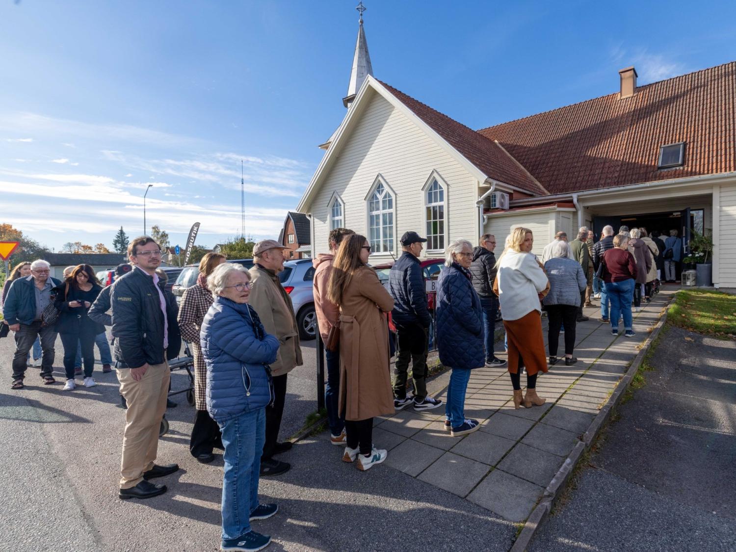 Människor står i en kö utanför en kyrkobyggnad en solig dag och väntar på att få komma in.