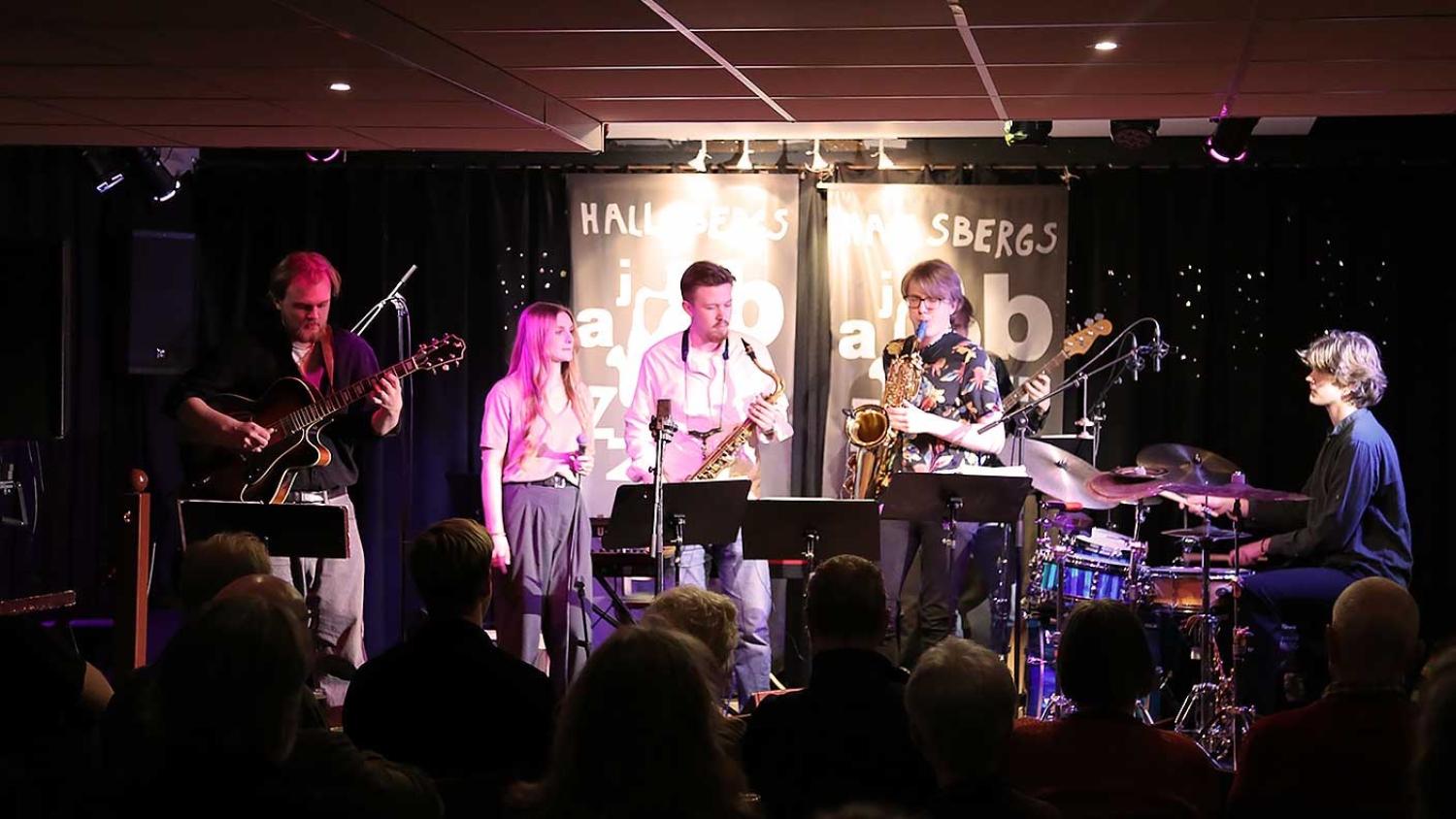 Fem musiker uppträder på scen med gitarr, saxofon, trummor och sång inför publik på en jazzklubb.
