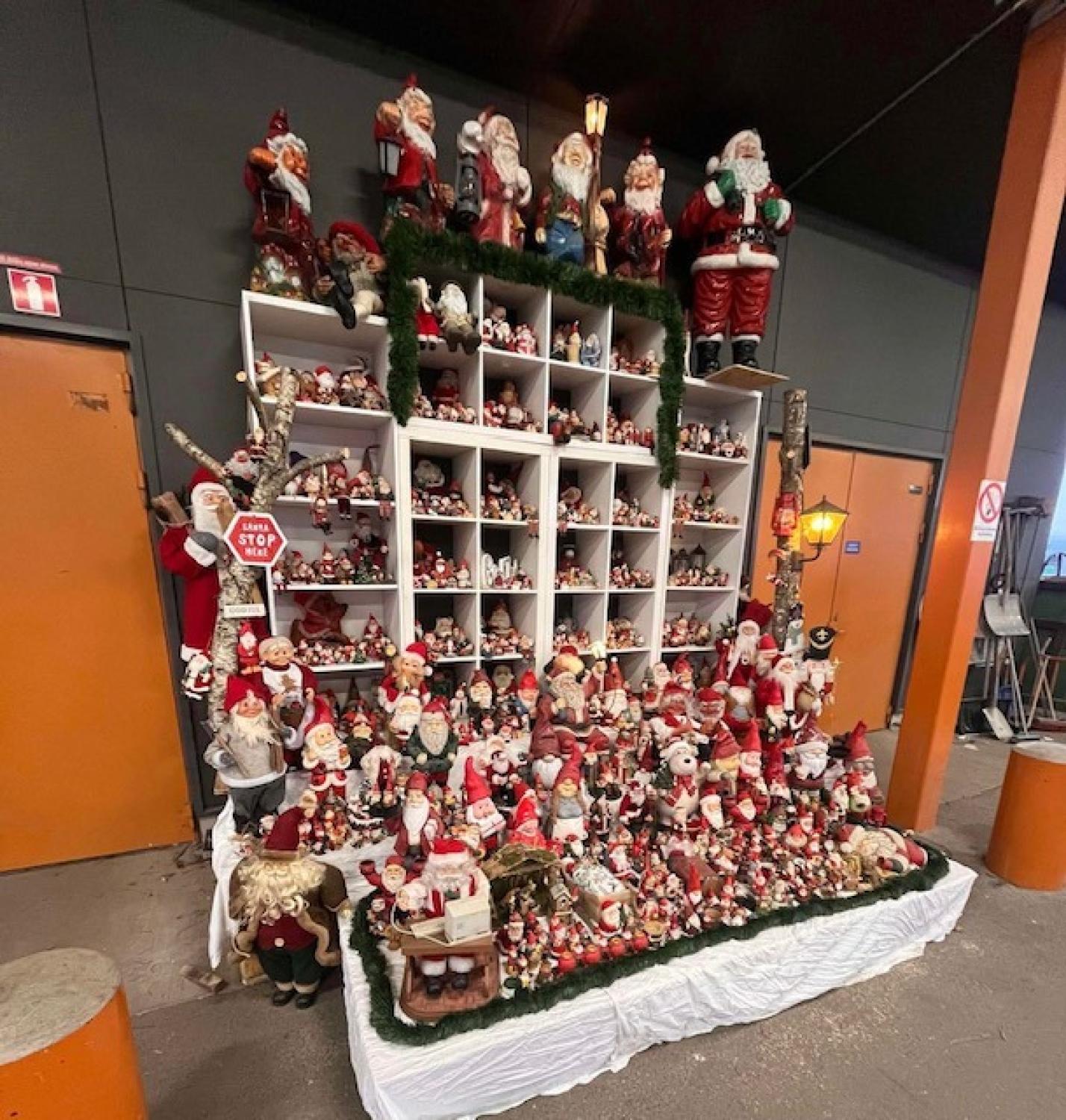 En stor utställning med olika tomtefigurer på vita hyllor och ett bord, placerade inomhus mot en orange vägg med dörrar.