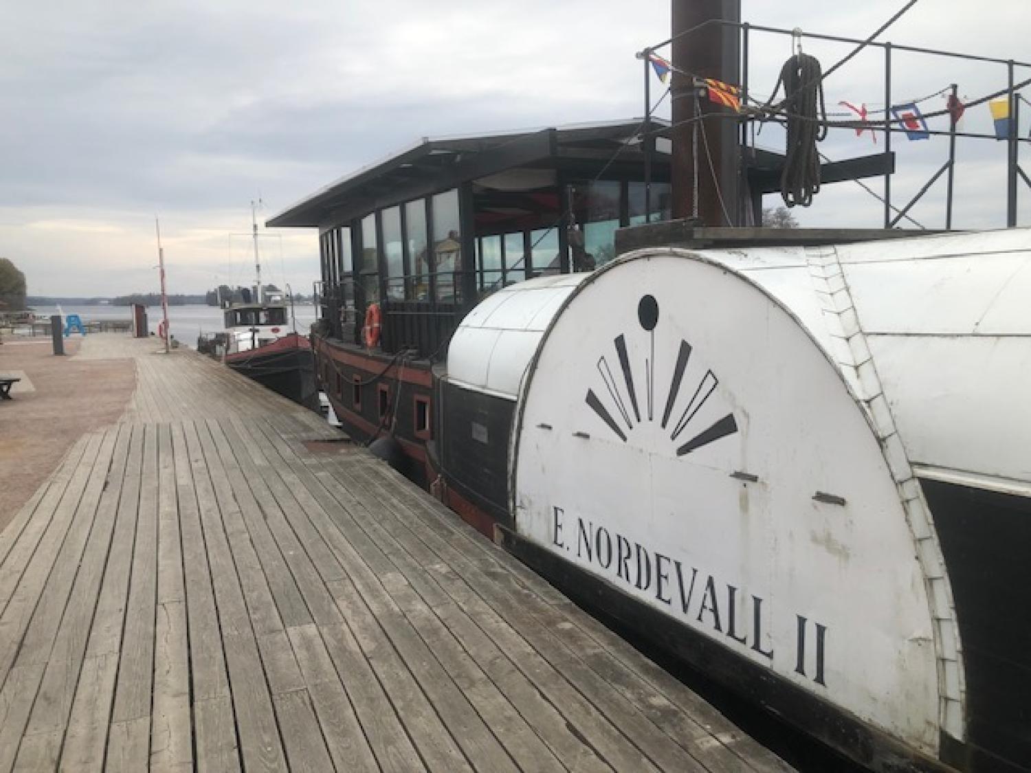 Ångbåten E. Nordevall II ligger vid en träbrygga med flaggor hängande ovanför och lugnt vatten i bakgrunden under en molnig himmel.