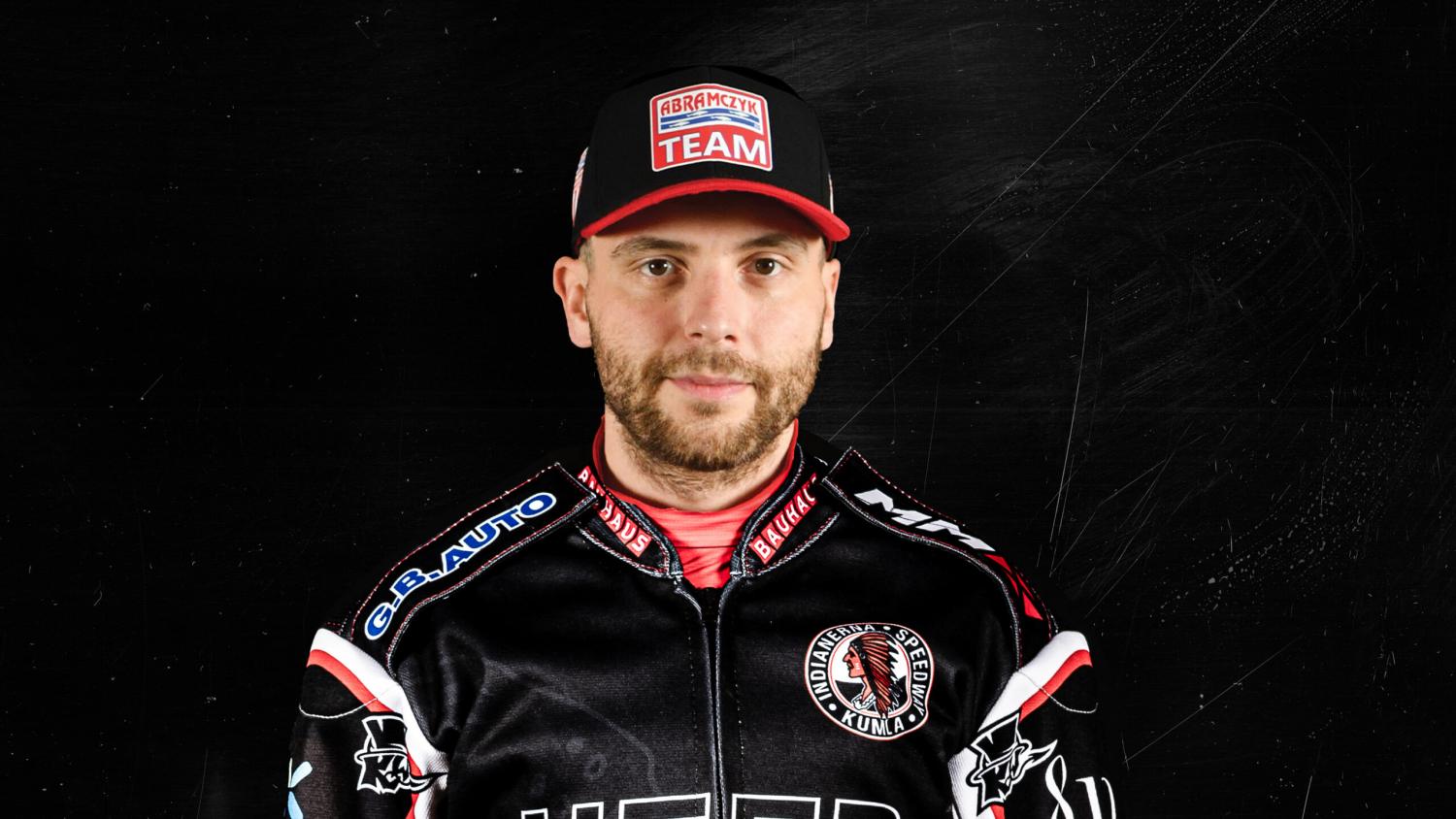 En man i svart racingdräkt med laglogotyper och en svart och röd keps står mot en svart bakgrund.