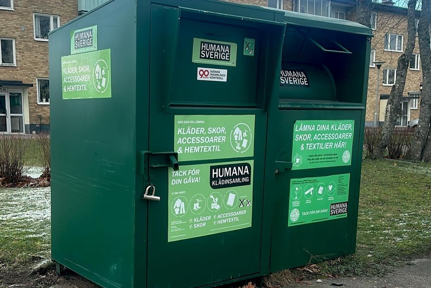 Två gröna Humana Sverige donationskärl för kläder, skor, accessoarer och textilier är placerade på en trottoar i närheten av flerfamiljshus.