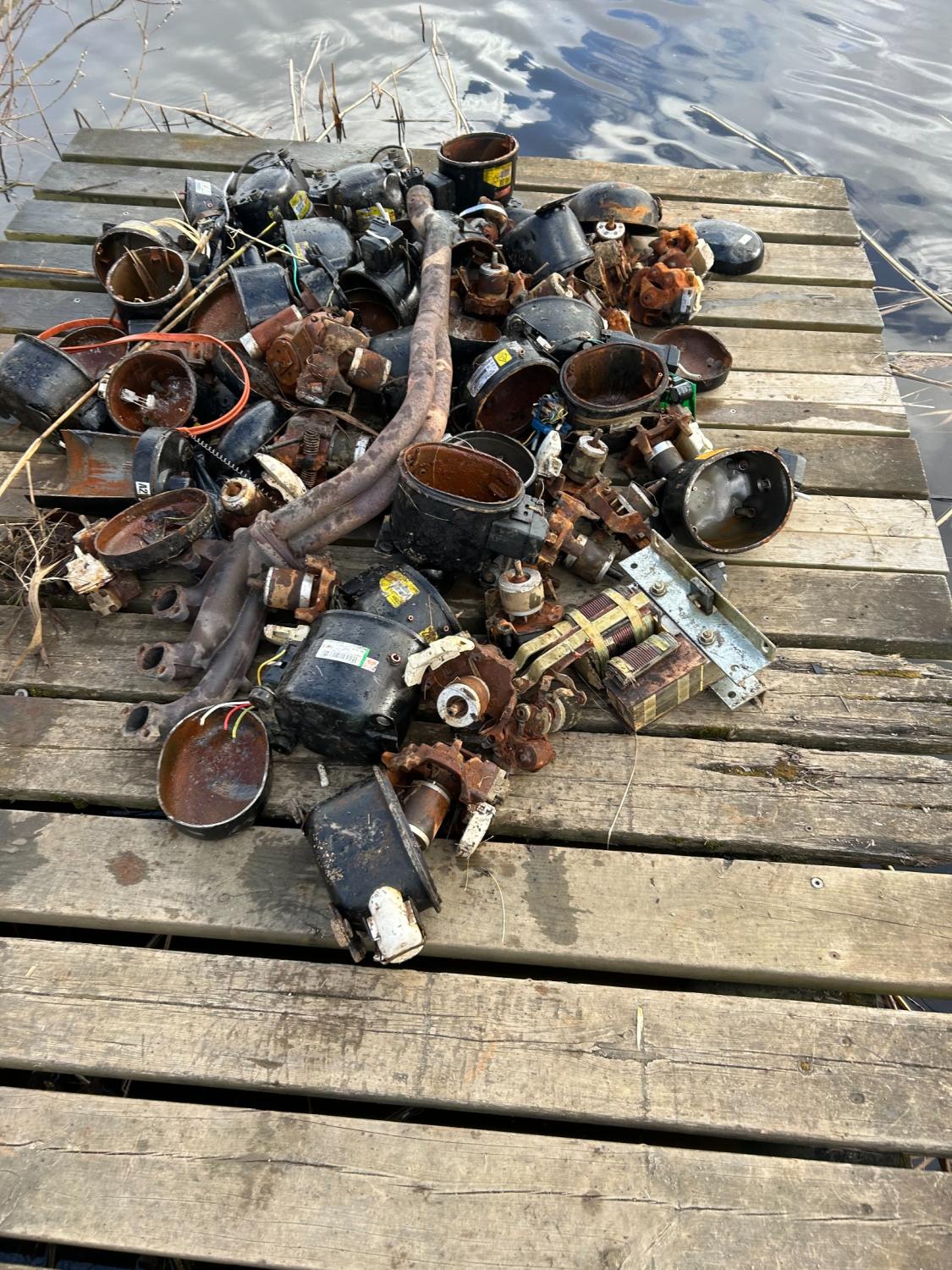 En hög med rostiga, trasiga rördelar och ventiler ligger på en träbrygga intill ett vattendrag.