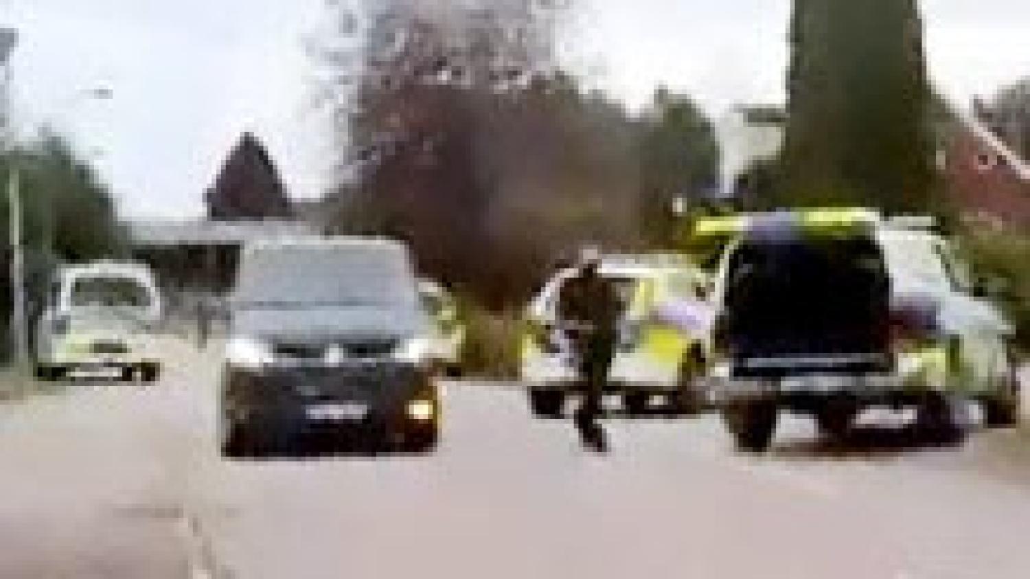 Flera polisbilar, en polis och en mörk bil står stilla på en gata i förorten med träd och hus i bakgrunden.