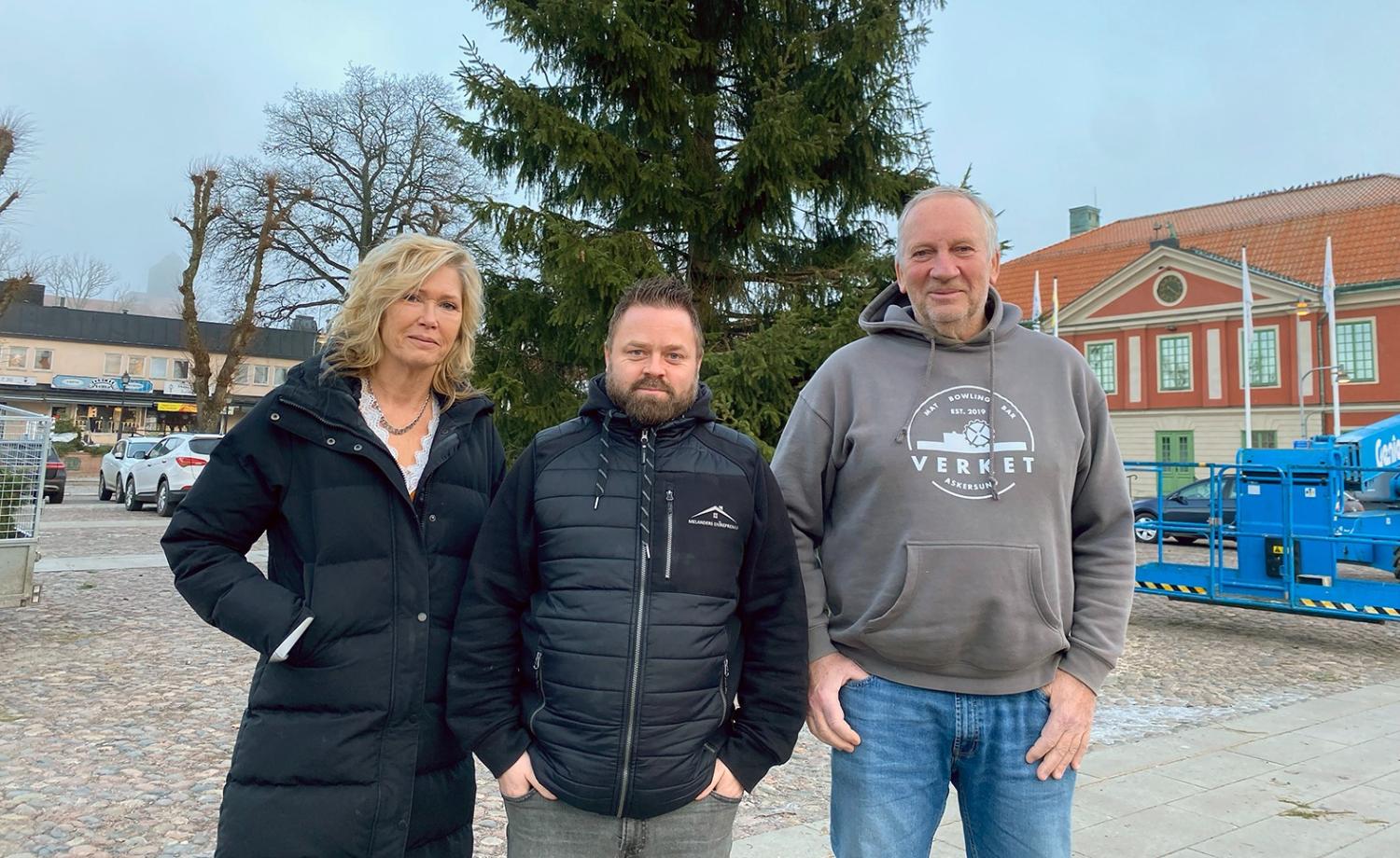 Tre vuxna står utomhus framför ett stort vintergrönt träd och byggnader, iklädda vinterjackor och fritidskläder.