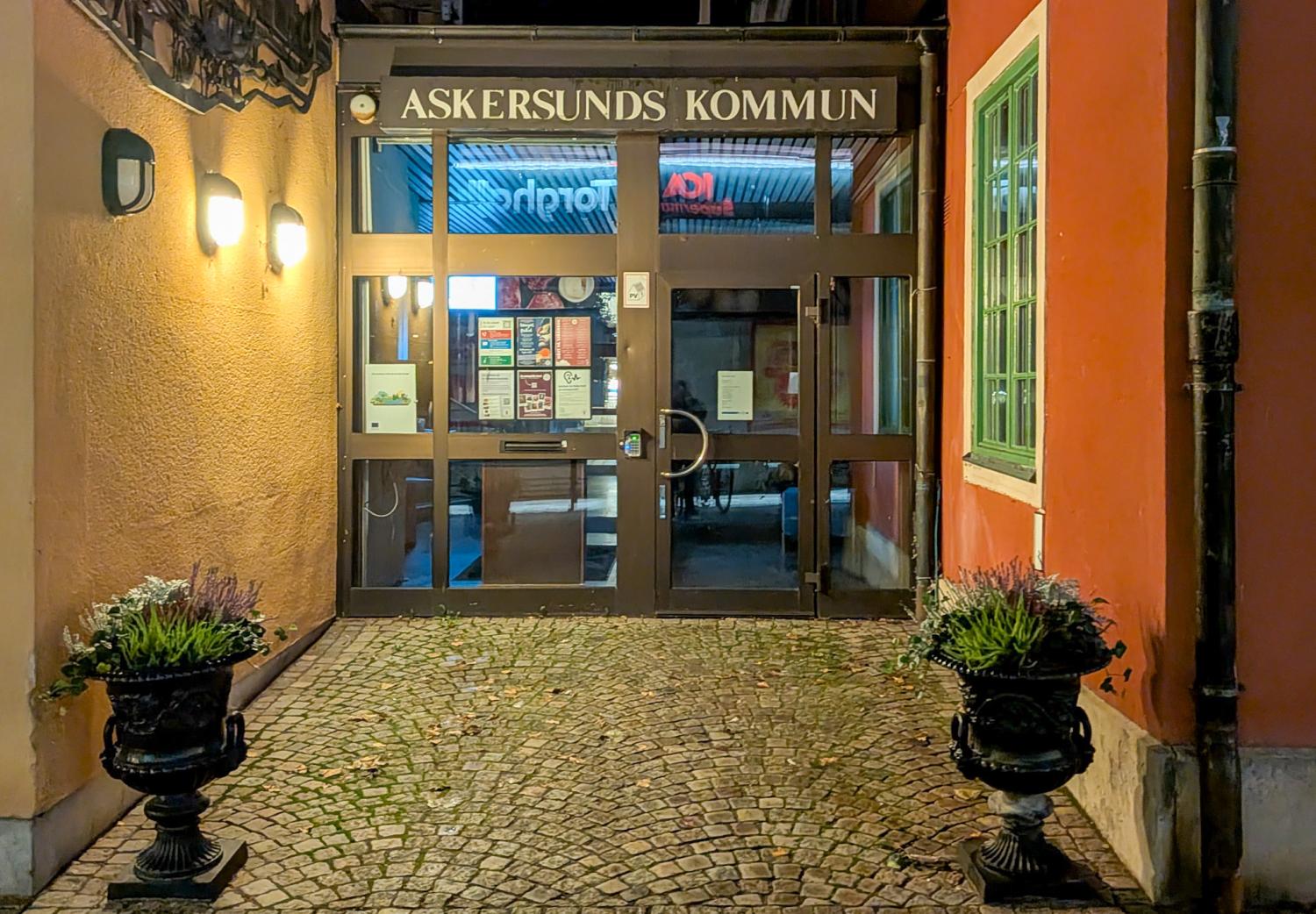 Ingången till Askersunds kommunhus på natten, med krukväxter på båda sidor om en kullerstensgång och upplysta vägglampor.