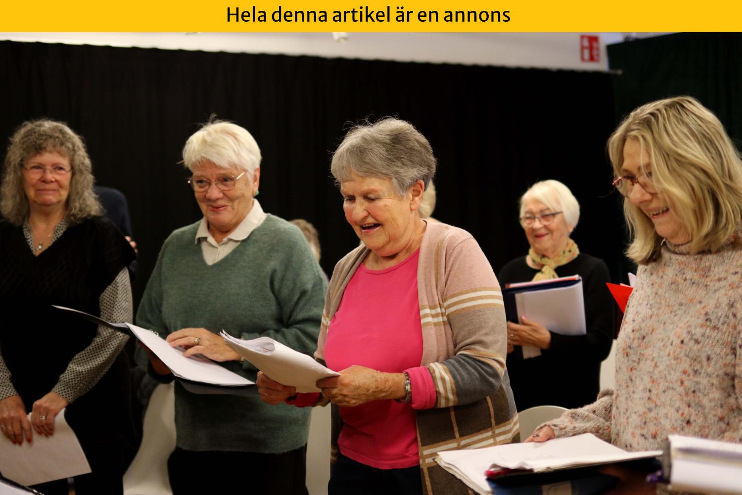 En grupp äldre vuxna står tillsammans inomhus, håller i papper och ler.