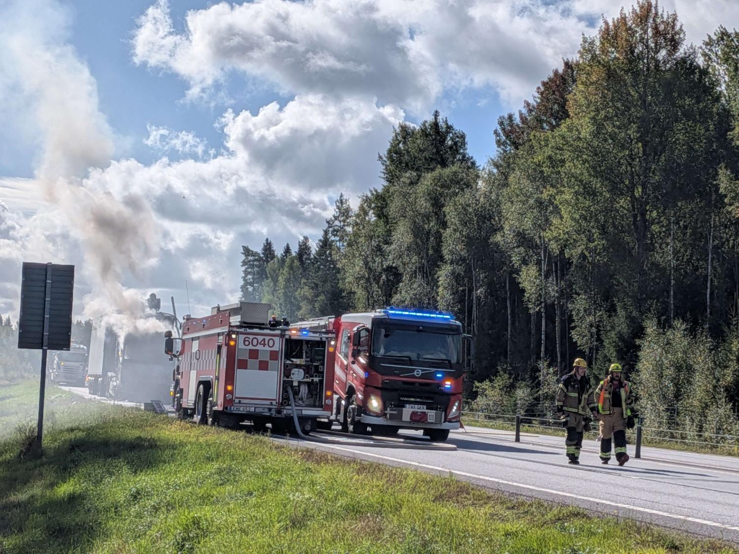 Två brandbilar med blinkande ljus står parkerade på en väg nära träden, medan rök stiger upp i bakgrunden och två brandmän går längs vägkanten.