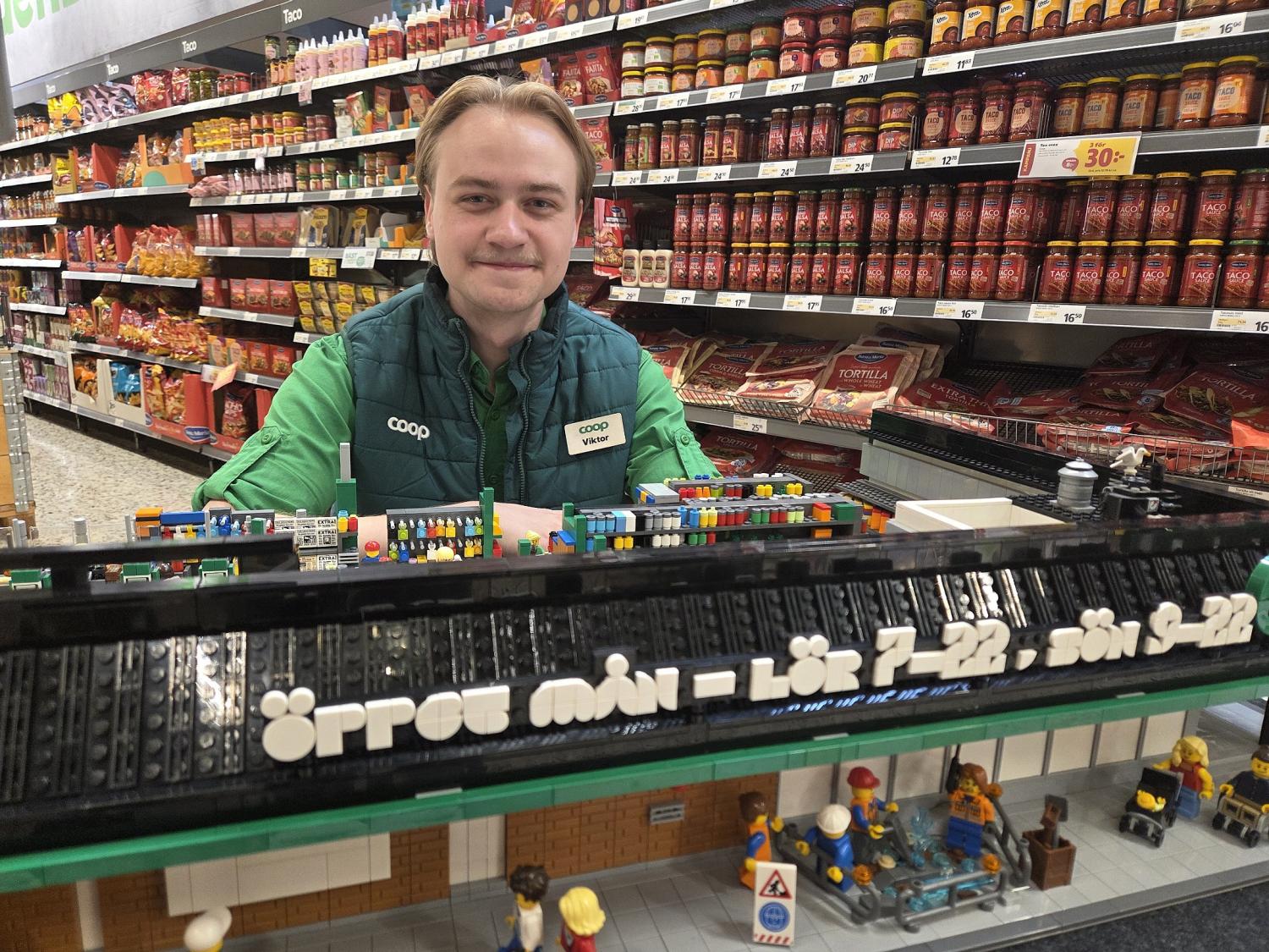 En butiksanställd står bakom en LEGO-modell av en stormarknad, där öppettiderna anges på svenska, med hyllor fyllda med varor i bakgrunden.