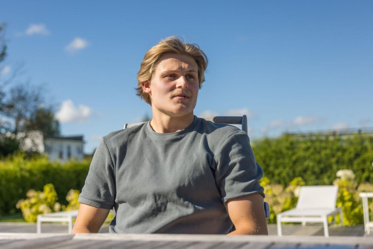 En ung man med blont hår sitter utomhus på en stol, iklädd en grå T-shirt, med gröna buskar och vita utemöbler i bakgrunden under en klarblå himmel.