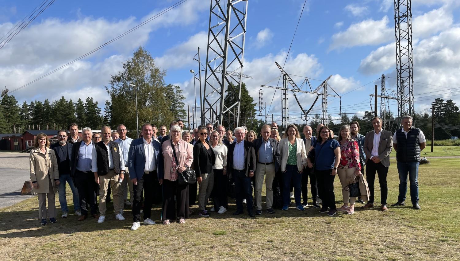 En grupp på trettio personer som poserar för ett foto utanför, står framför elektriska metalltorn och utrustning med träd och en delvis molnig himmel i bakgrunden.