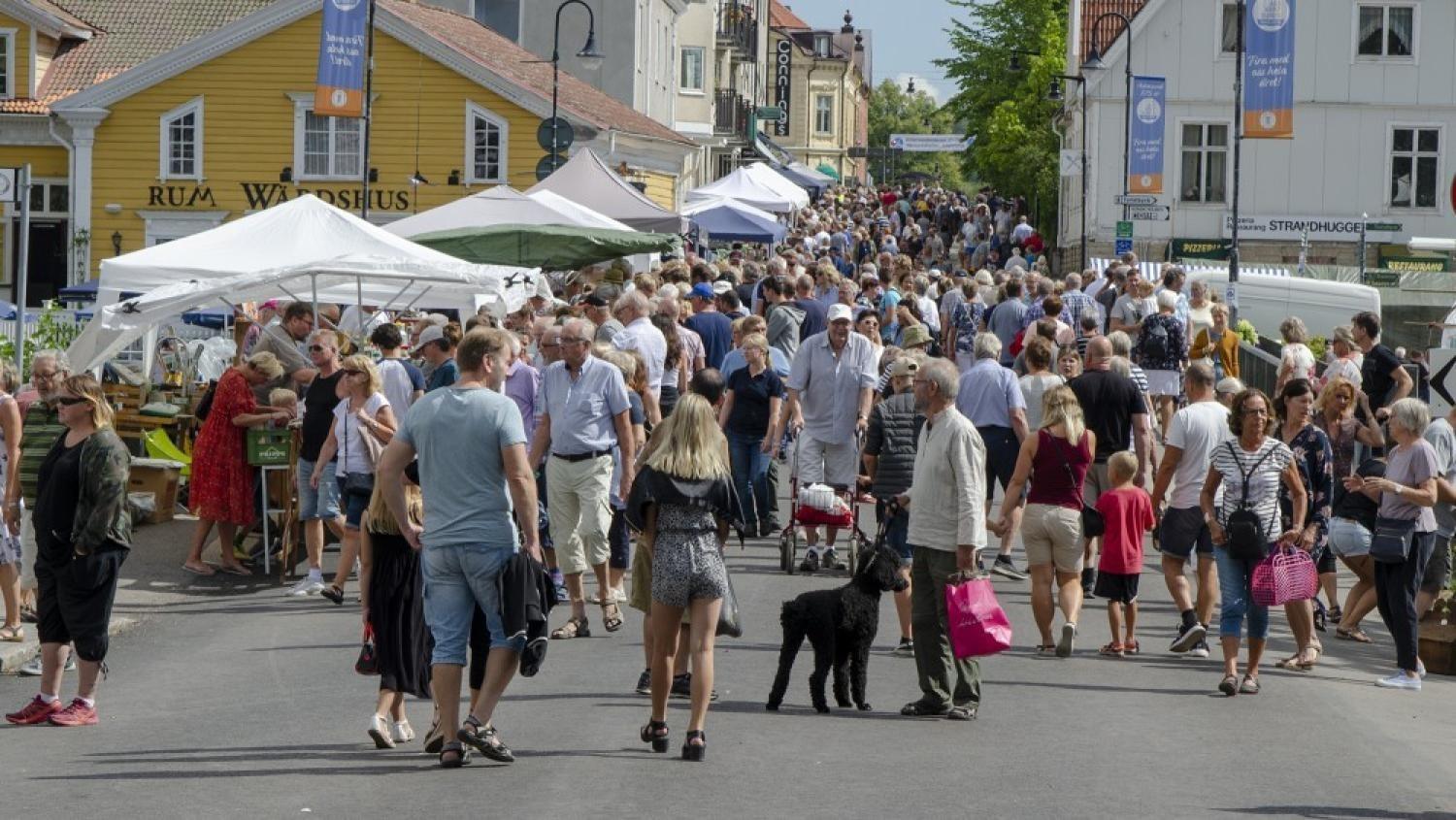 En folkmassa som promenerar och handlar på en utomhusmarknad, med försäljartält, byggnader och skyltar synliga i bakgrunden.