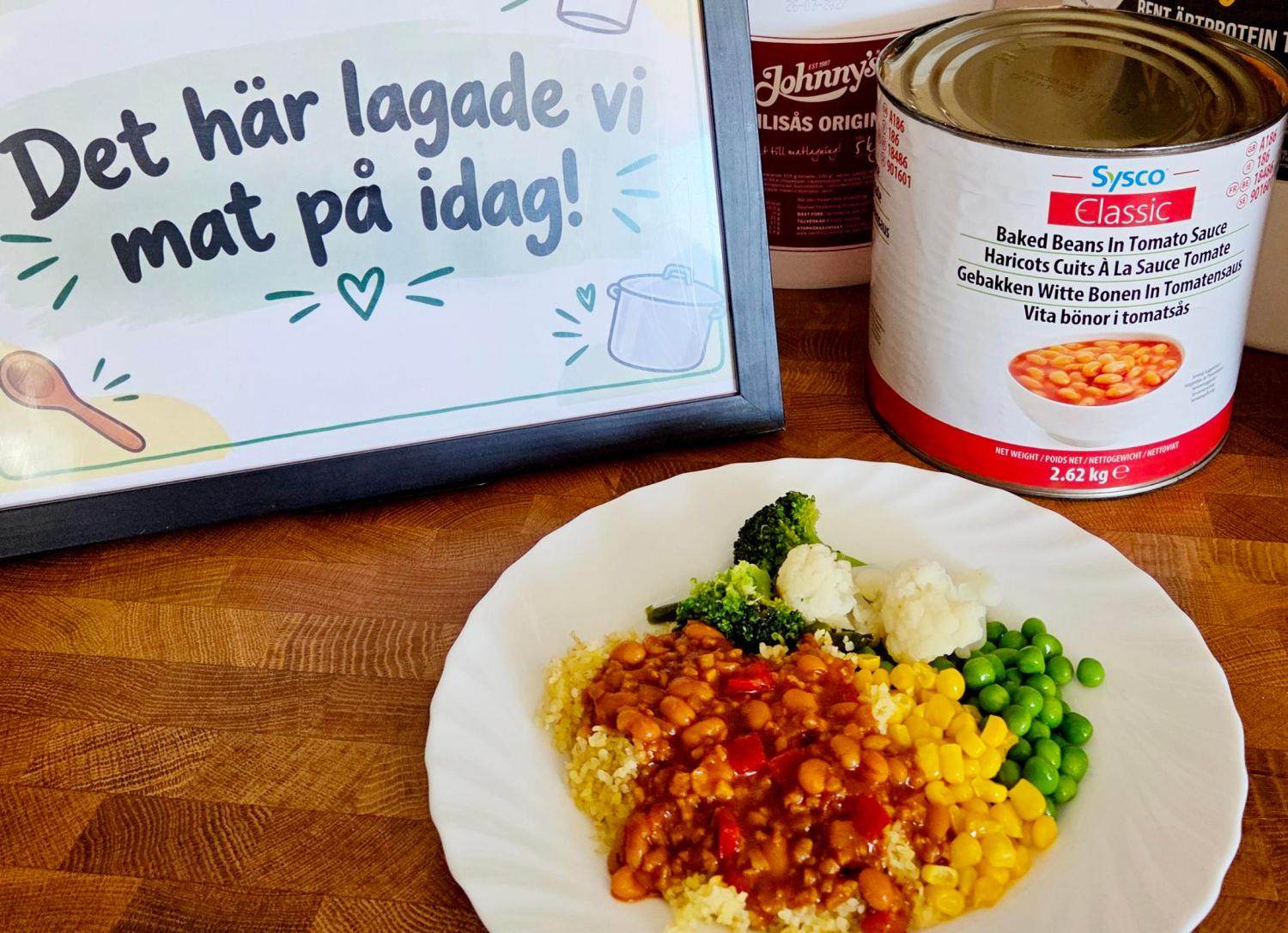 En tallrik med bakade bönor, majs, ärtor, broccoli och blomkål står bredvid en burk bönor och en skylt på svenska där det står: ”Det här lagade vi mat på idag!”.