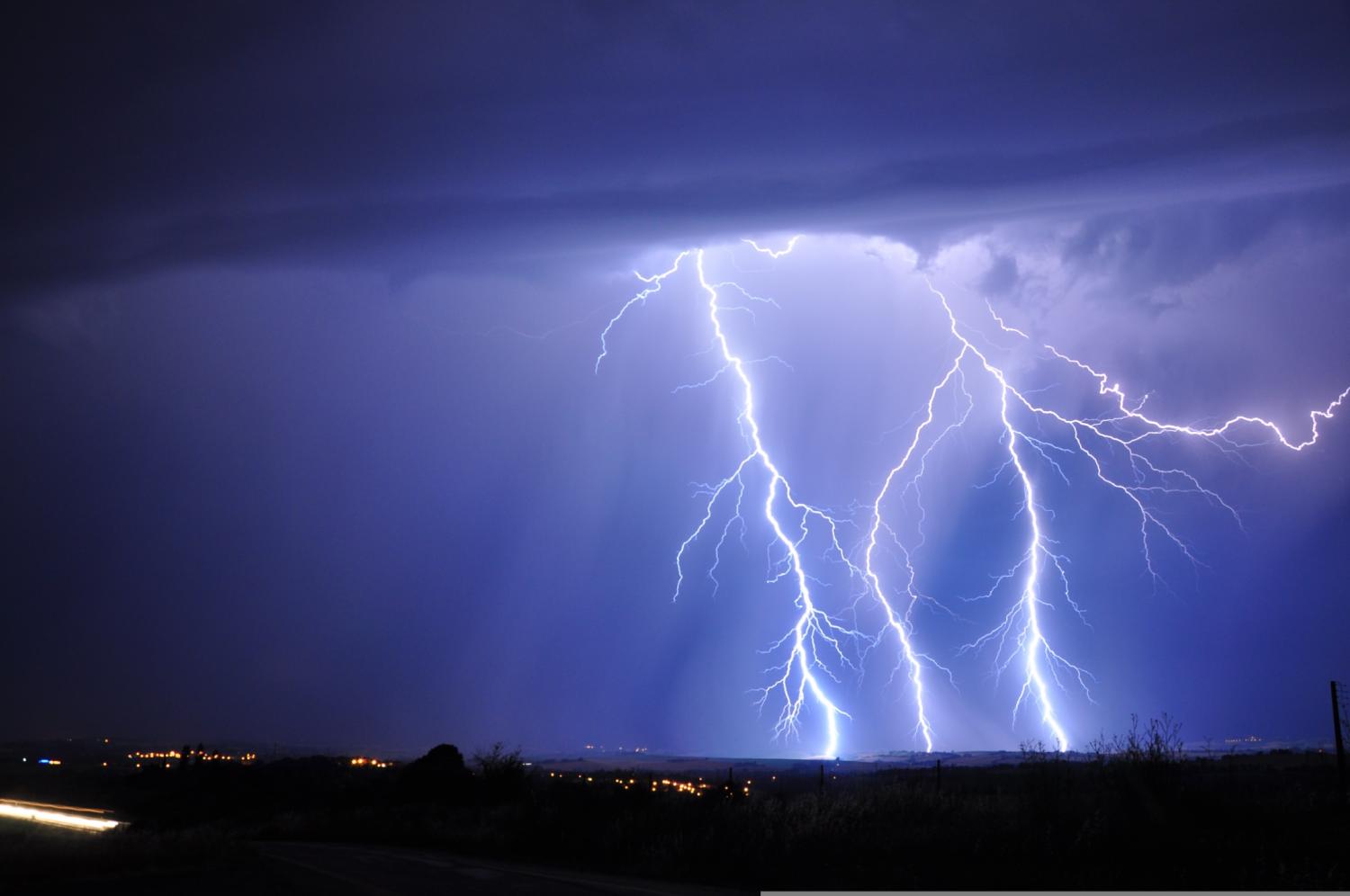 Flera blixtar slår ner i marken under mörka stormmoln på natten, och stadens ljus syns vid horisonten.
