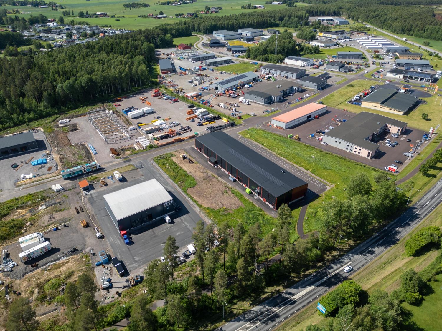 Flygfoto över ett industriområde med flera lagerlokaler, kontor, parkerade fordon, byggarbetsplatser samt omgivande grönområden och vägar.