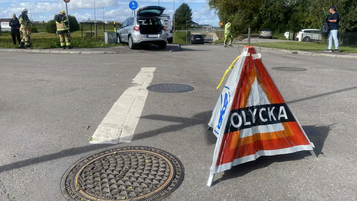 En vägskylt med texten ”Olycka” står uppställd intill en öppen bagagelucka, och i bakgrunden syns räddningspersonal.