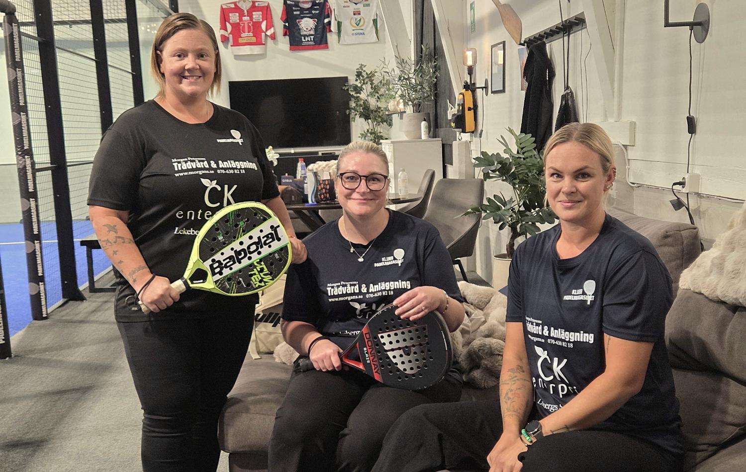 Tre kvinnor i svarta tröjor sitter och står inomhus med padelracketar i händerna, medan sportutrustning och tröjor syns i bakgrunden.
