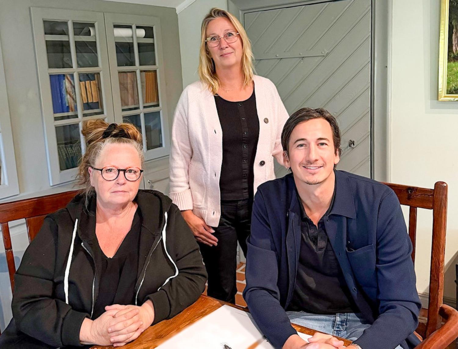 Tre vuxna, två sittande och en stående bakom dem, poserar inomhus vid ett träbord, och alla tittar mot kameran.