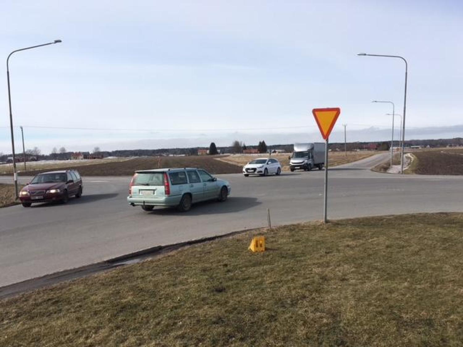 Fyra bilar närmar sig och navigerar i en rondell på landsbygden, med en avkörningsskylt synlig i förgrunden och fält i bakgrunden.