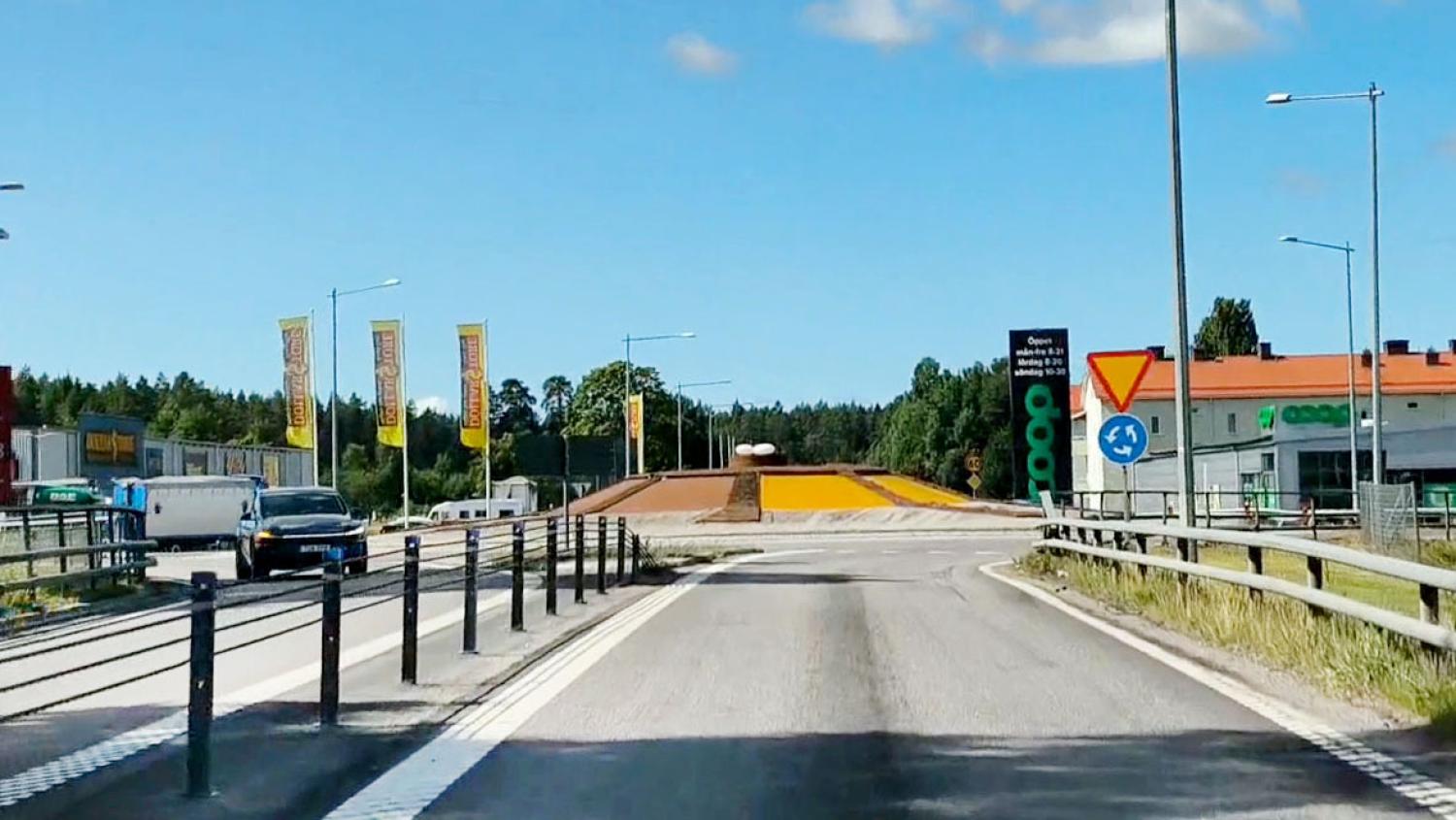 En väg leder fram till en rondell med bilar, en gräsbevuxen kulle i mitten, trafikskyltar och byggnader i närheten, allt under en klarblå himmel.