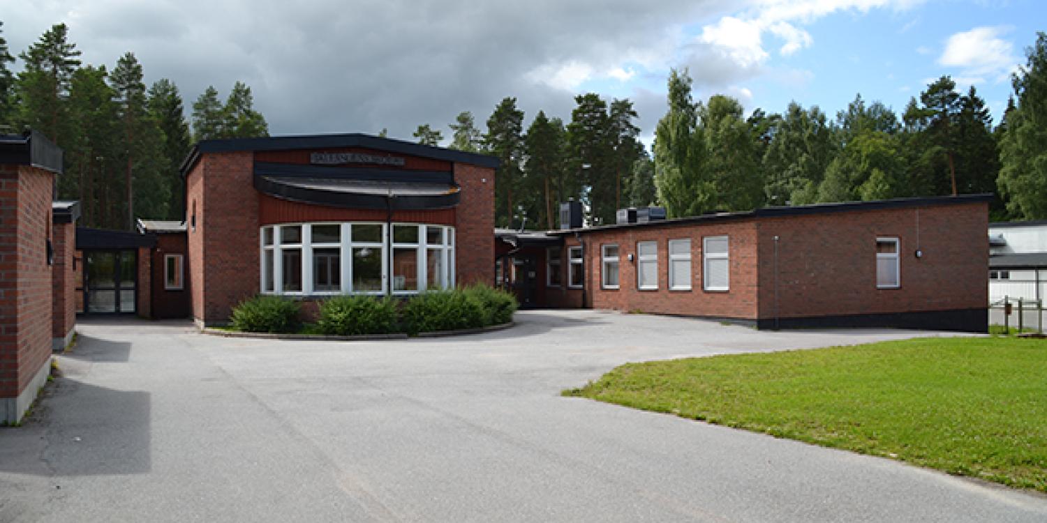 En skolbyggnad i tegel med stora fönster och platt tak, omgiven av träd och en asfalterad innergård under en molnig himmel.
