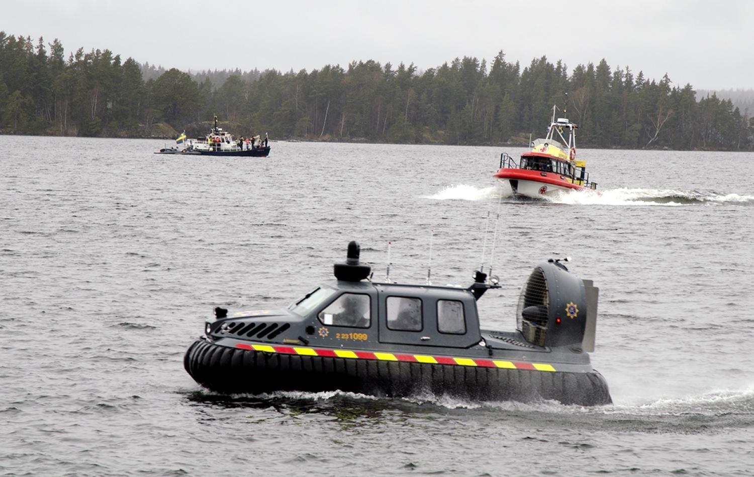 En svävare och två räddningsbåtar färdas över en sjö med en skogsklädd strandlinje i bakgrunden under en molnig himmel.