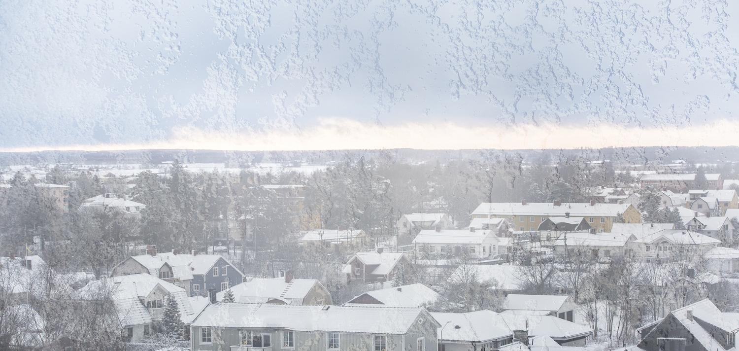 Ett snölandskap sett genom ett frosttäckt fönster, med förortshus och träd täckta i vitt.