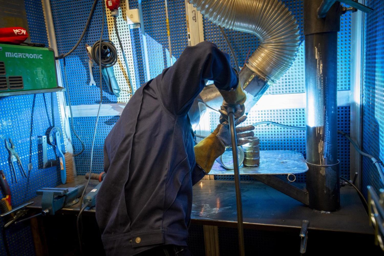 En person iförd handskar och skyddsjacka svetsar metall vid en arbetsbänk, med ventilation och verktyg synliga i bakgrunden.