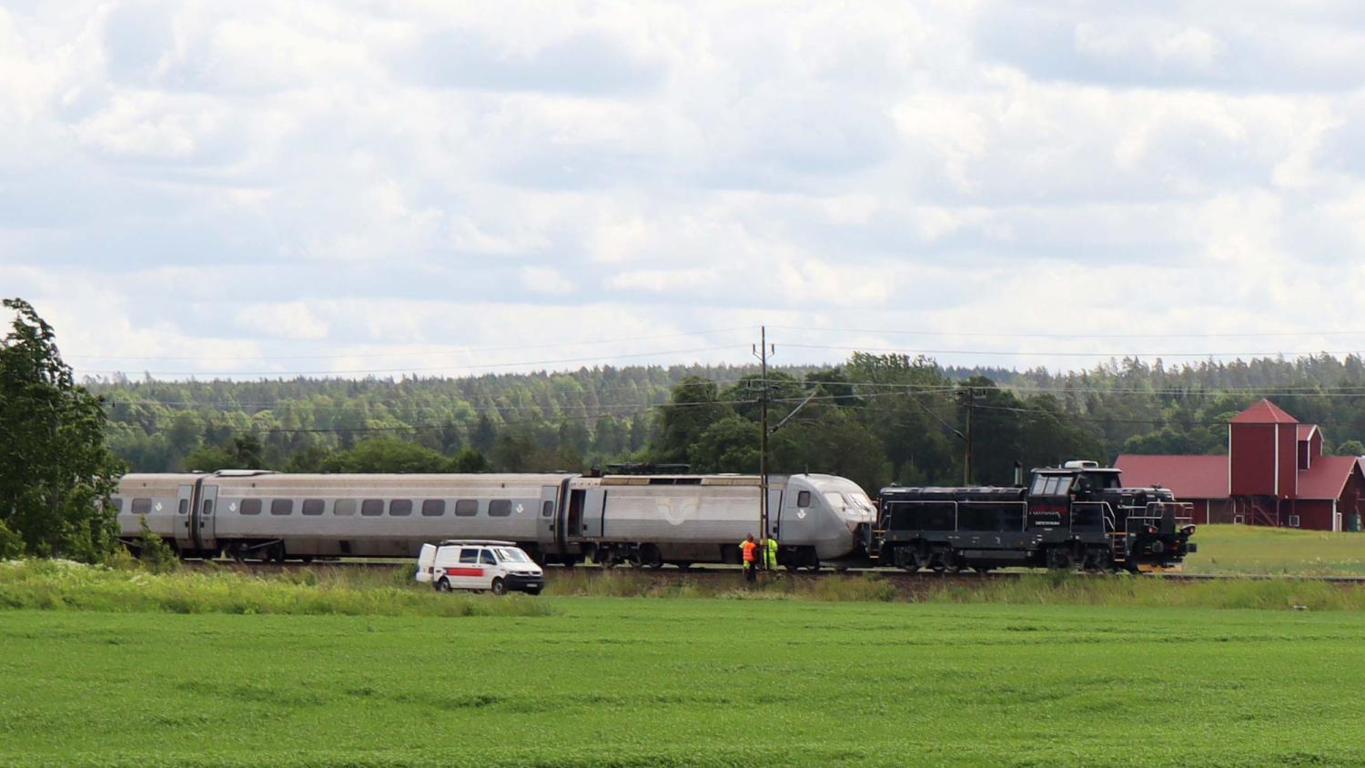 Ett tåg och två underhållsfordon står stilla på spåren i ett landsbygdsområde med gröna fält, träd och en röd lada i bakgrunden. En vit skåpbil och tre personer befinner sig i närheten.