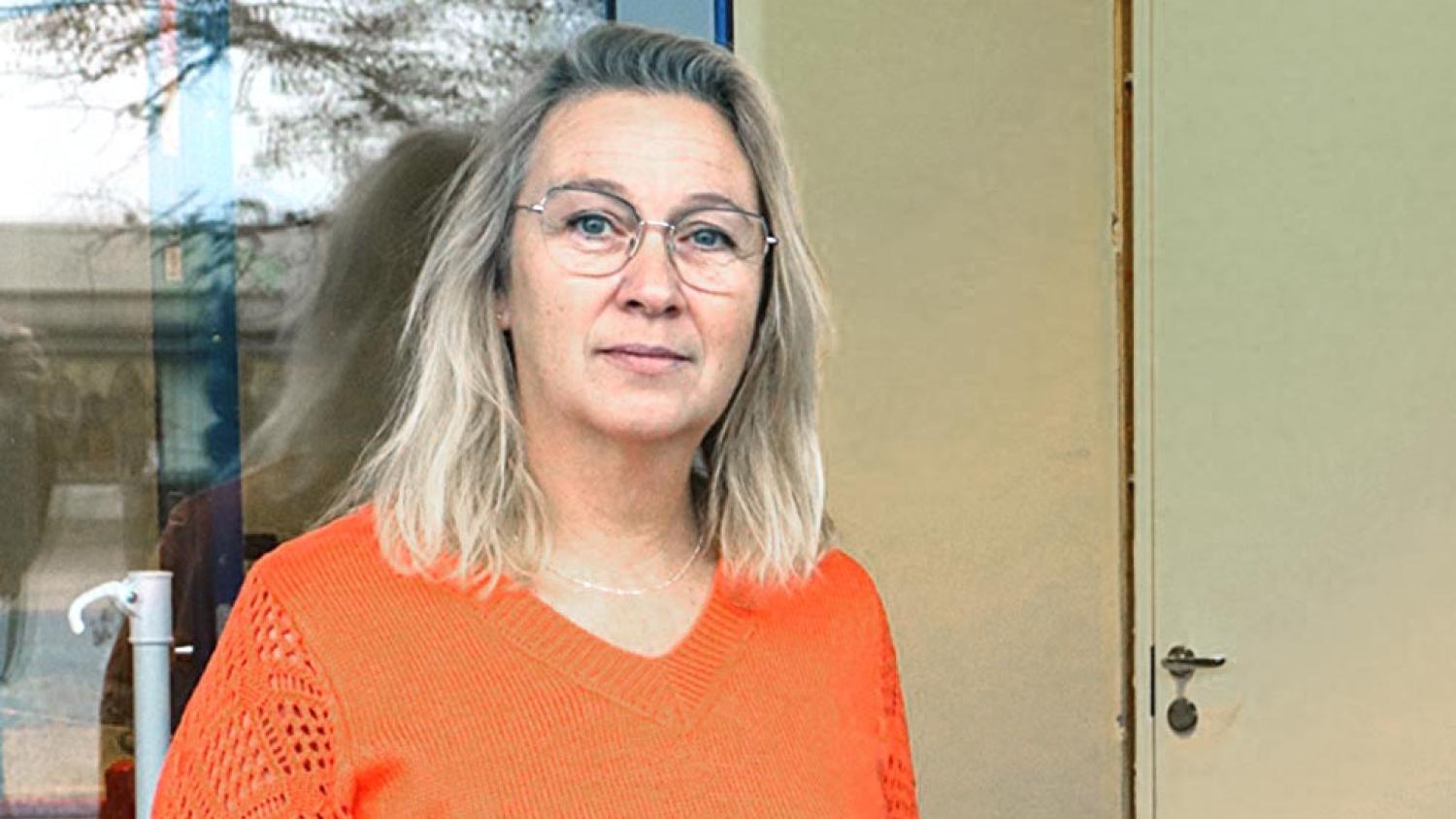 En kvinna med blont hår och glasögon, klädd i en orange tröja, står inomhus framför en beige dörr och ett fönster.