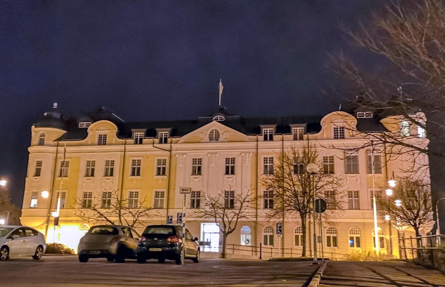 En stor, upplyst byggnad med välvda fönster och mörkt tak syns på natten, med parkerade bilar och lövlösa träd framför.