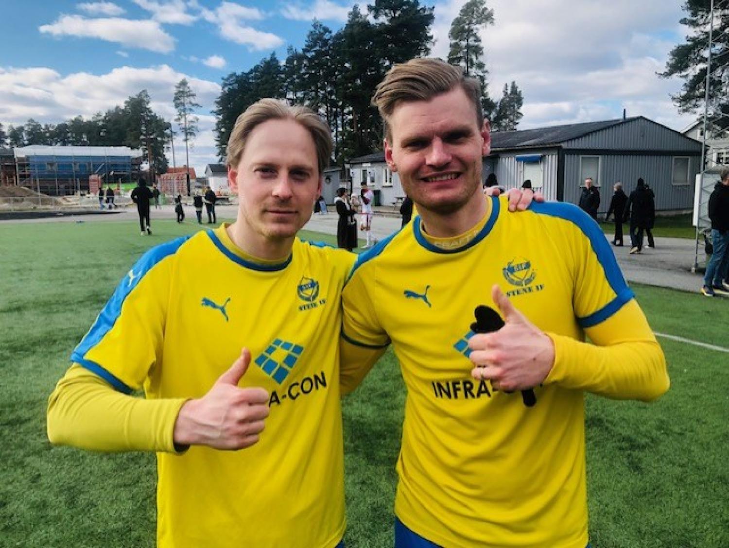 Två fotbollsspelare i gula tröjor med blå accenter gör tummen upp på en gräsplan, med träd och byggnader i bakgrunden.