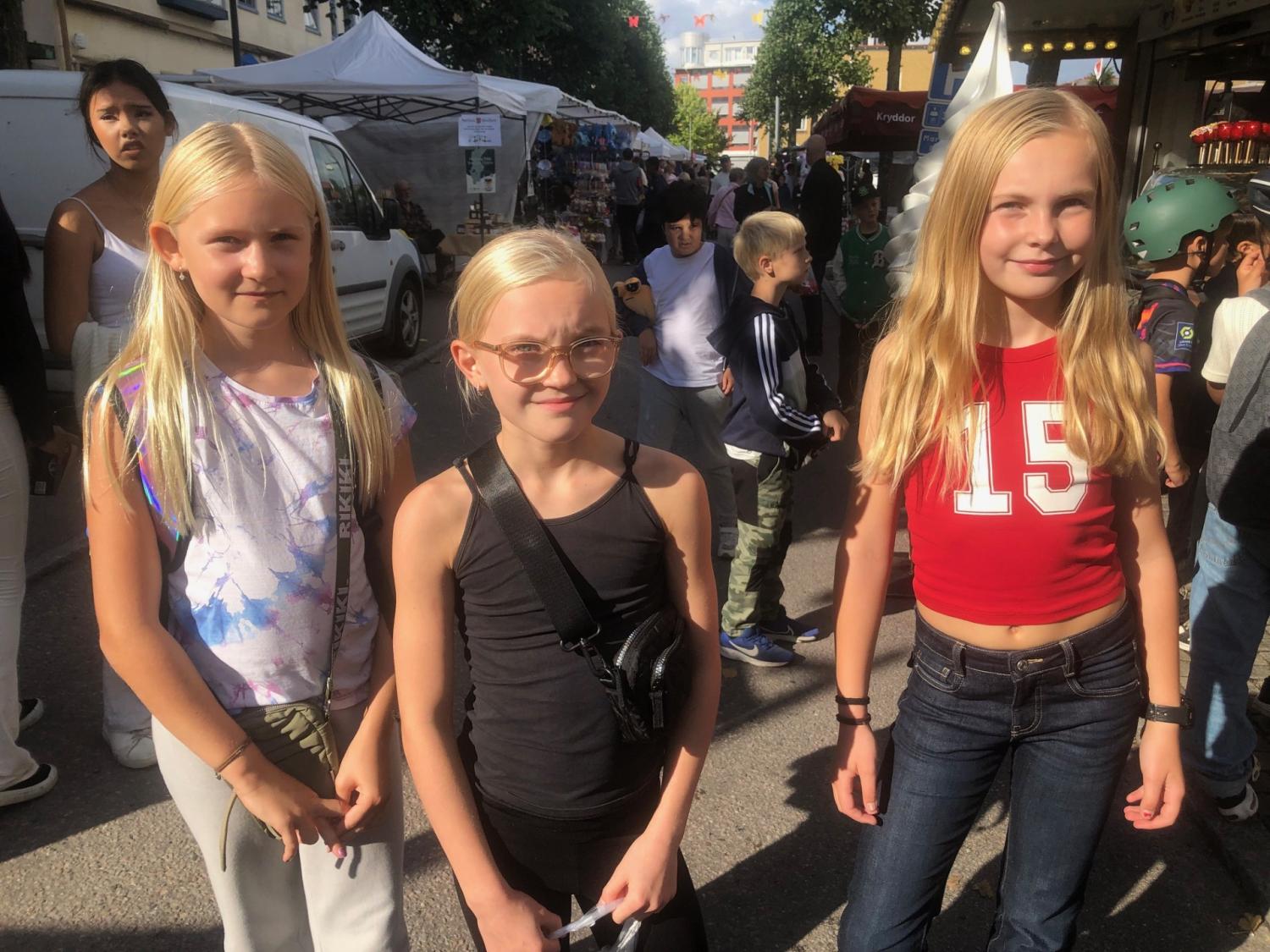 Tre unga flickor med blont hår står tillsammans utomhus på en gatumarknad, med andra människor och stånd synliga i bakgrunden en solig dag.