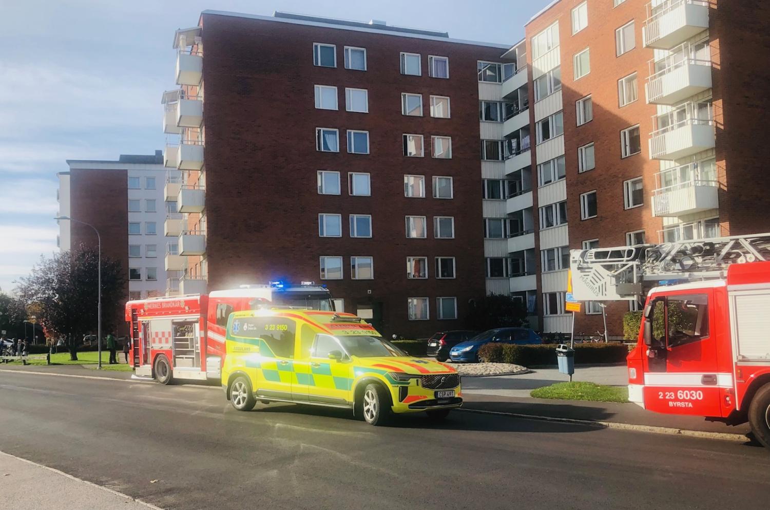 En ambulans och två brandbilar står parkerade utanför ett flervåningshus en klar dag.