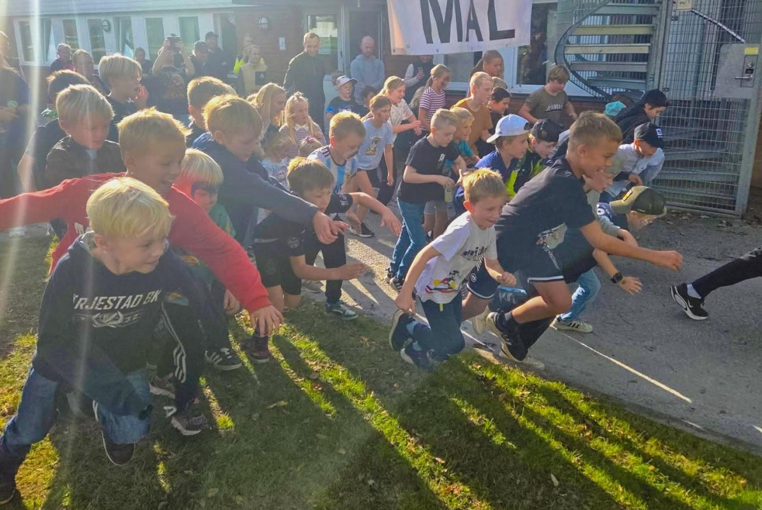 En grupp barn springer mot en mållinje utomhus, med vuxna som tittar på och en banderoll med texten