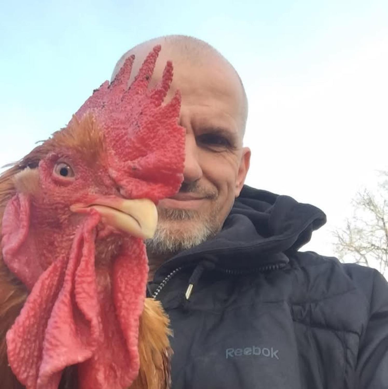 En man i svart jacka står utomhus och håller en tupp nära sitt ansikte så att deras ögon är i höjd med varandra, vilket skapar en jämförelse sida vid sida.