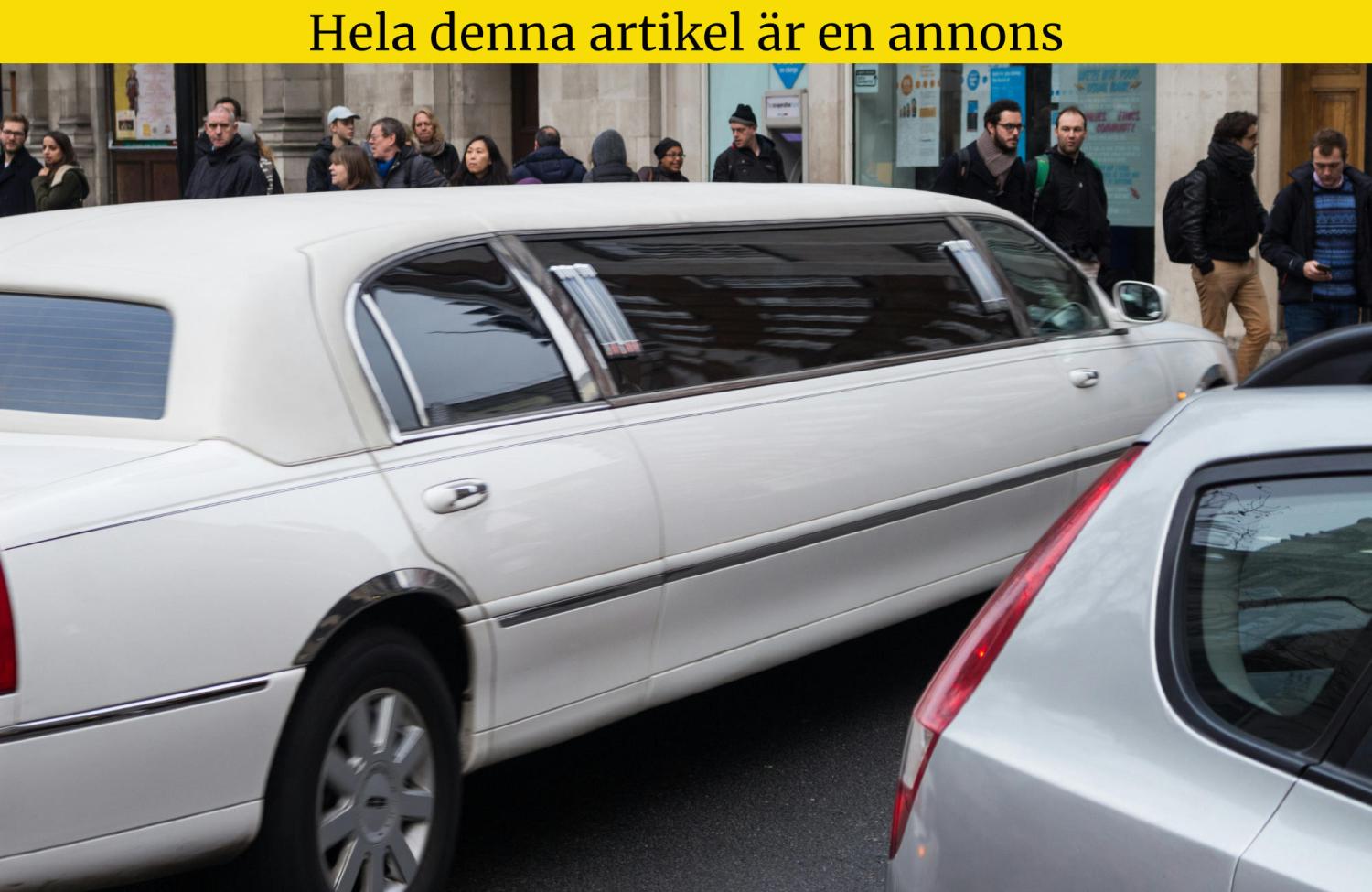 En vit limousine kör nerför en livlig gata bredvid fotgängare och parkerade bilar.