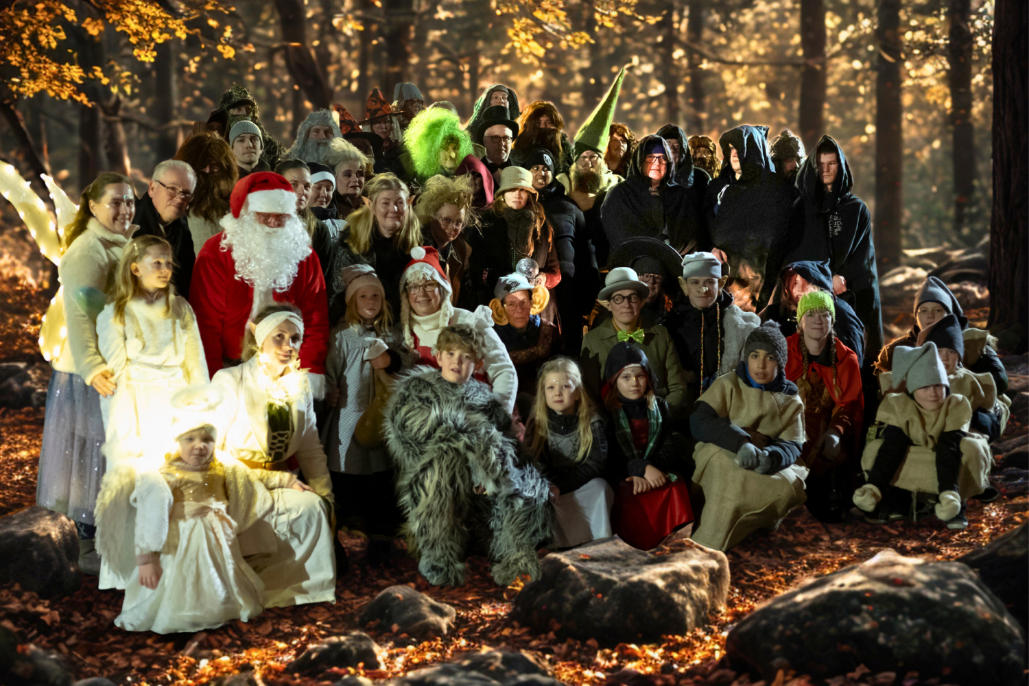 En stor grupp människor klädda i olika fantasi- och djurkostymer poserar tillsammans i en solbelyst skog med höstlöv på marken.