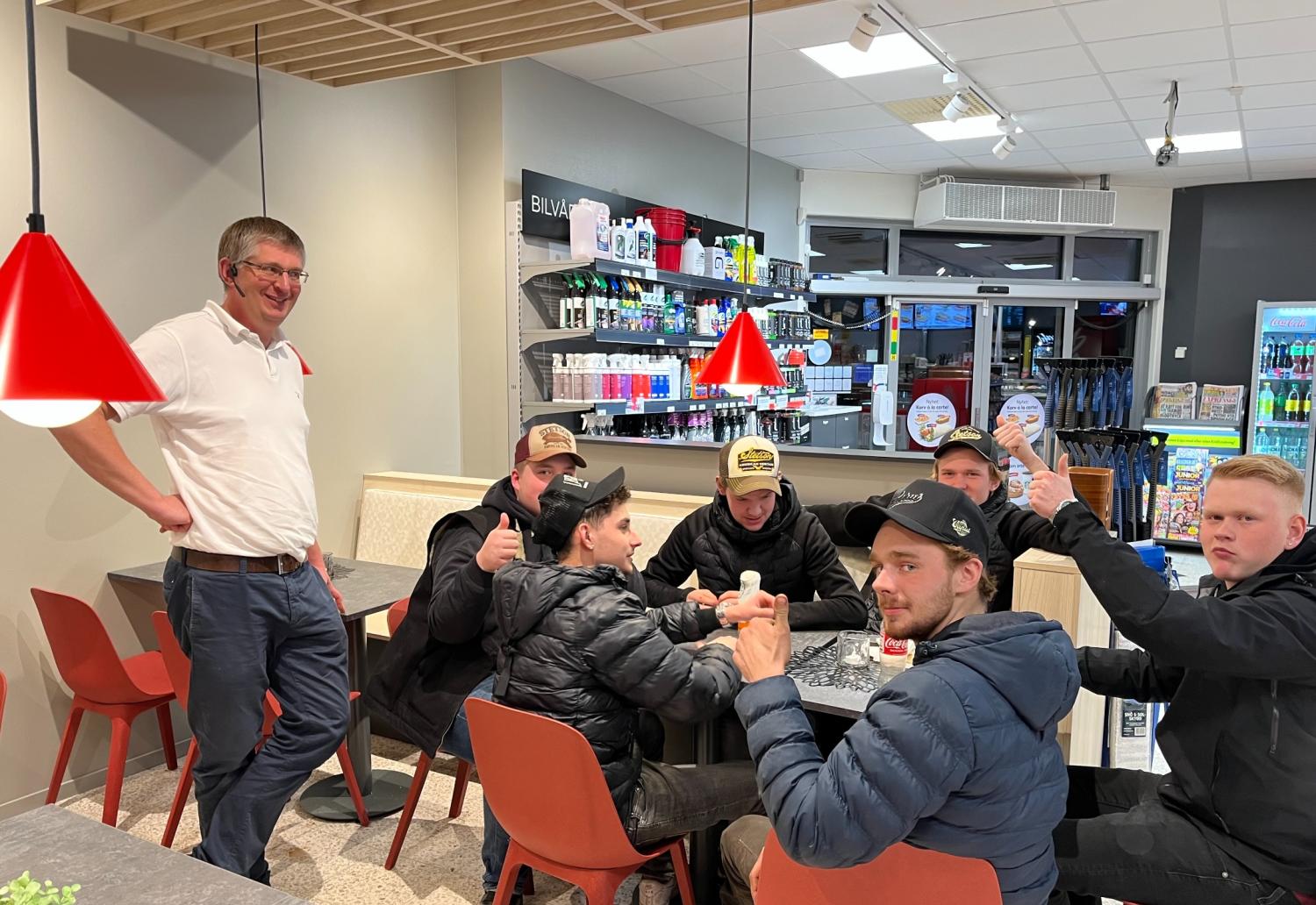 En grupp på sex unga män sitter runt ett bord i en butik eller på ett café, några gör tummen upp, medan en äldre man står bredvid och ler. Hyllor med produkter syns i bakgrunden.