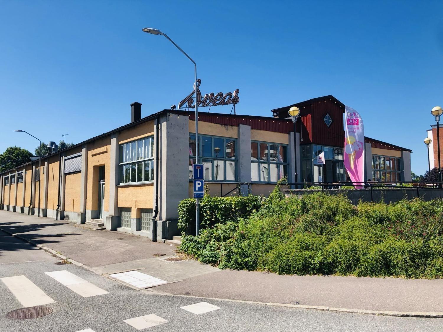 En stor byggnad i industriell stil med gula tegelväggar, stora fönster och en metallskylt på taket, belägen vid ett soligt gathörn med grönska och en flagga.