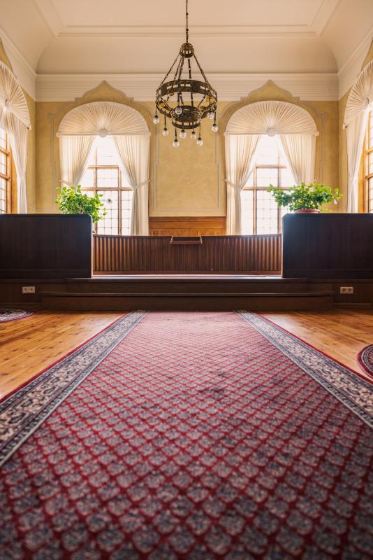 Interiör i en traditionell synagoga med träbänkar, en central röd matta och fönster som flankerar en utsmyckad ljuskrona.
