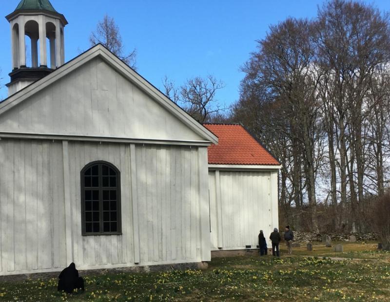 Flera personer är utspridda på området nära en liten vit träkyrka med rött tegeltak, omgiven av träd och gräs med gula blommor.