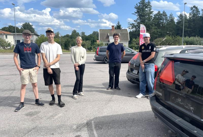 Fem personer står på avstånd från varandra på en parkeringsplats en solig dag, med bilar och träd i bakgrunden.