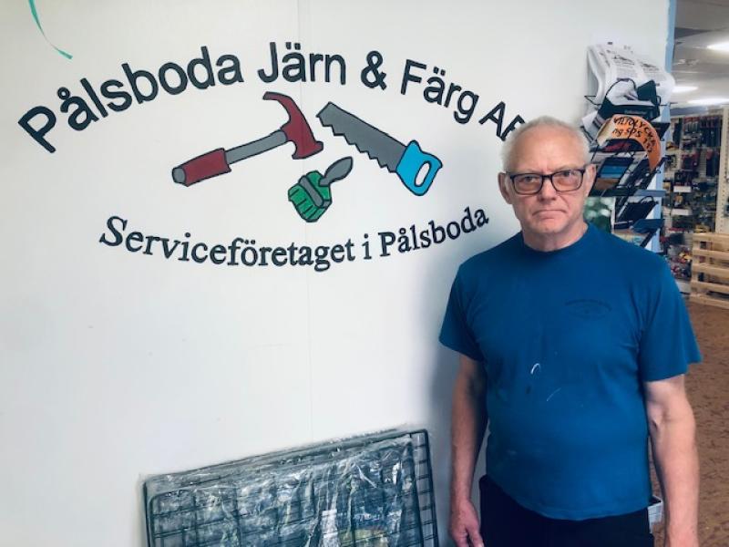 En äldre man i glasögon och blå skjorta står intill en vägg med logotypen för Pålsboda Järn & Färg AB, med verktyg och svensk text.