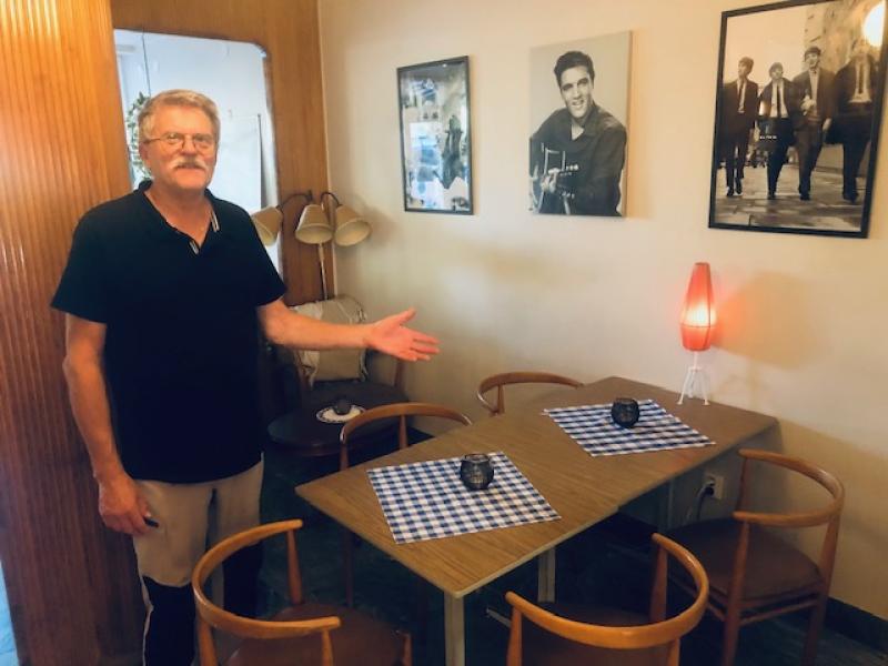 En man står vid ett litet matbord med fyra trästolar, rutiga bordstabletter och inramade foton på väggen i ett mysigt rum.
