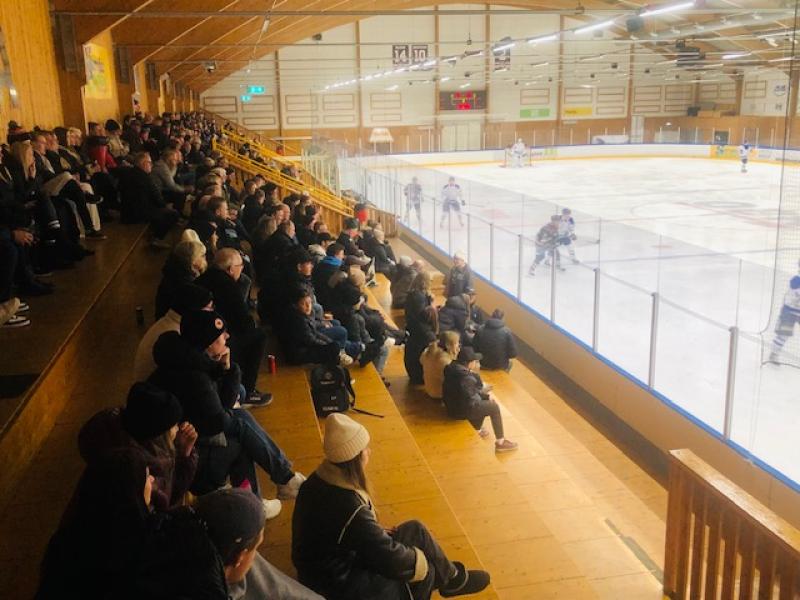 Publiken sitter på en träläktare och tittar på en ishockeymatch som pågår i en inomhusarena.