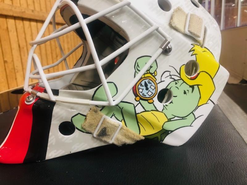 En målvaktsmask för hockey med en illustration av en grön tecknad sköldpadda som bär en gul hatt och håller i ett fickur.