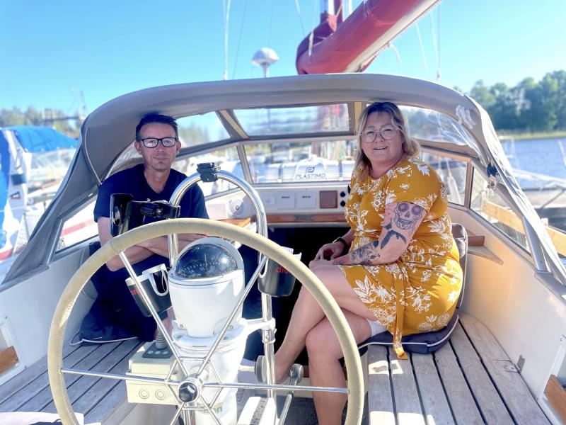 Två personer sitter i sittbrunnen på en segelbåt, med ratten och navigationsutrustningen framför sig. En person är klädd i en svart outfit och den andra är i en gul blommig klänning.