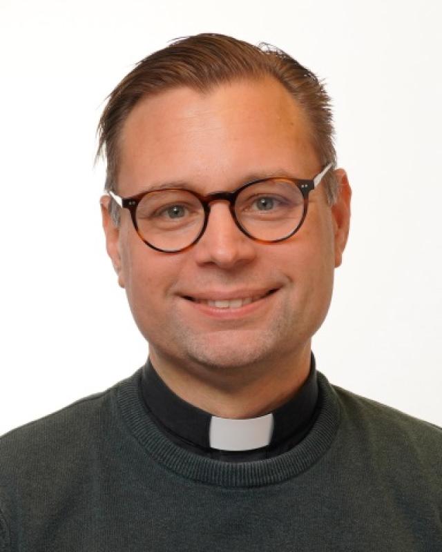 En man med kort brunt hår och glasögon, klädd i prästkrage och mörk tröja, ler mot en vit bakgrund.