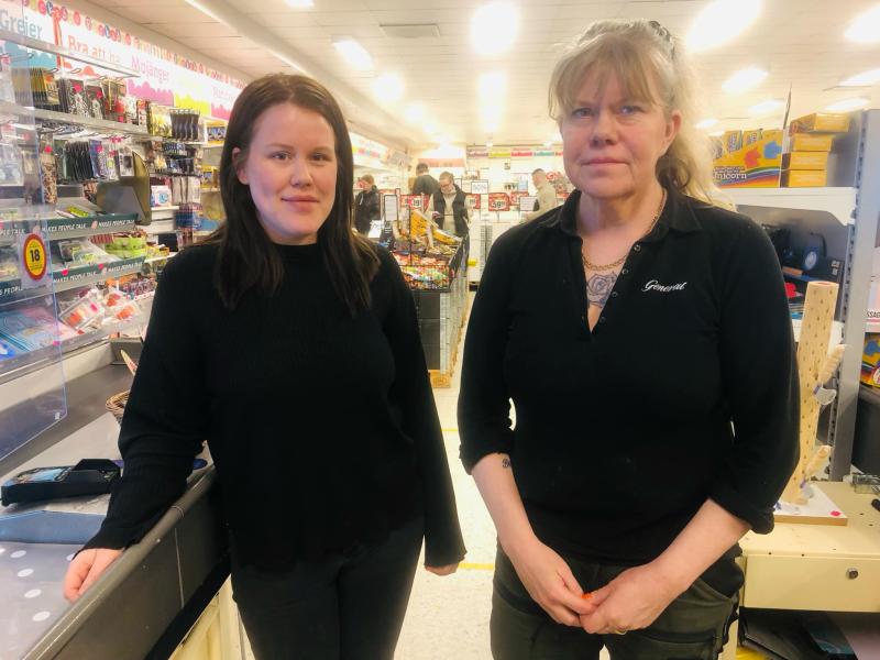 Två kvinnor står inne i en butik nära kassan. Hyllor med olika produkter syns i bakgrunden.
